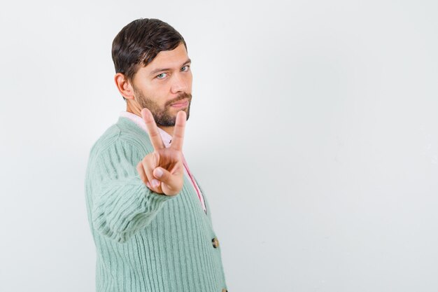 Jeune homme en chemise, cardigan montrant un geste de paix et ayant l'air confiant.