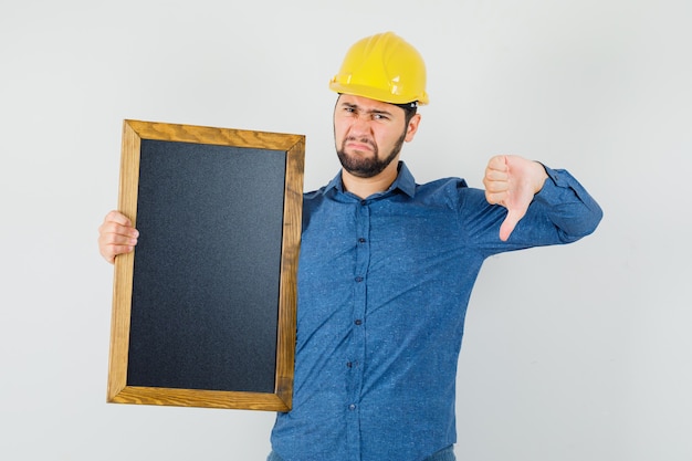 Jeune homme en chemise, casque tenant tableau noir, montrant le pouce vers le bas et l'air mécontent, vue de face.