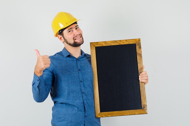 Jeune homme en chemise, casque tenant tableau noir, montrant le pouce vers le haut et regardant gai, vue de face.