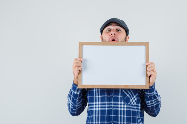 Jeune homme en chemise, casquette tenant un cadre vide et regardant étonné, vue de face.