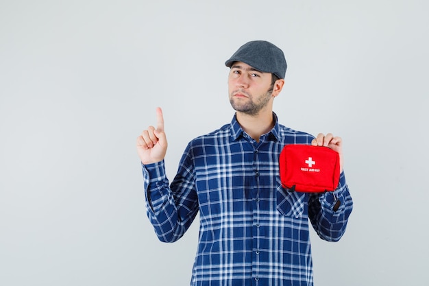 Jeune homme en chemise, casquette tenant une trousse de premiers soins, pointant vers le haut et regardant pensif, vue de face.