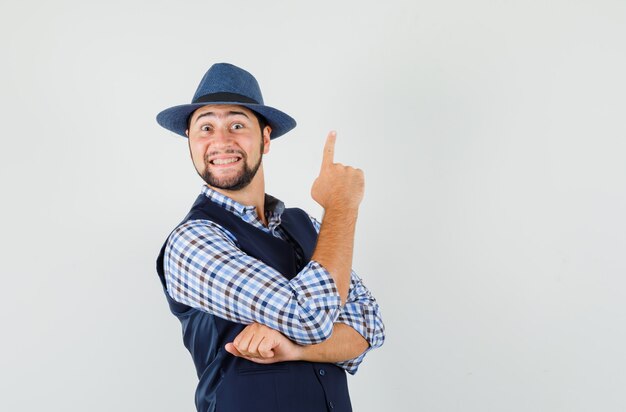 Jeune homme en chemise, gilet, chapeau pointant le doigt vers le haut et à la joyeuse vue de face.