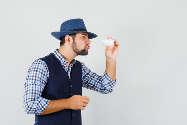 Jeune homme en chemise, gilet, chapeau à la recherche en bouteille de pilules et à la curieuse, vue de face.