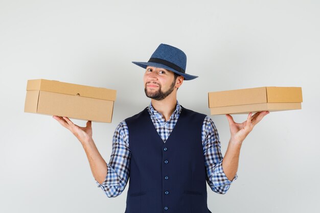 Jeune homme en chemise, gilet, chapeau tenant des boîtes en carton et à la vue de face, confiant.