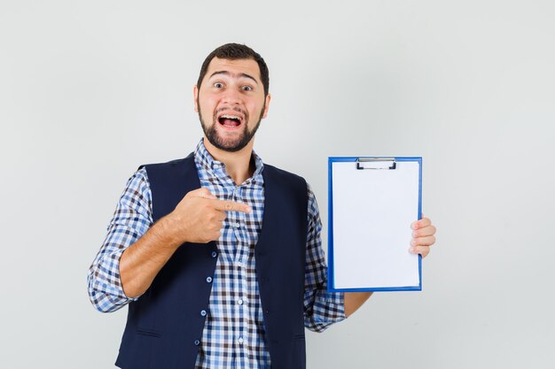 Jeune homme en chemise, gilet pointant sur le presse-papiers et à la joyeuse vue de face.