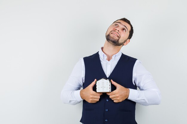 Jeune homme en chemise, gilet tenant le modèle de maison et à la recherche d'espoir