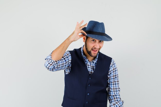 Jeune homme en chemise, gilet touchant son chapeau avec les doigts et à la recherche élégante, vue de face.