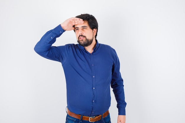 Jeune homme en chemise, jeans en détournant les yeux tout en tenant la main sur le front et en ayant l'air confiant, vue de face.