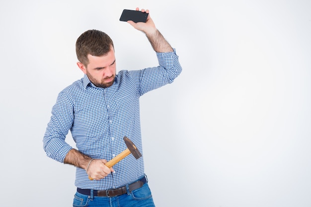 Jeune homme en chemise, jeans faisant semblant de jeter le téléphone portable tout en gardant le marteau et à la sérieuse, vue de face.