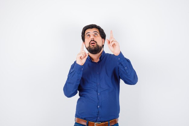 Jeune homme en chemise, jeans pointant vers le haut et l'air étonné, vue de face.