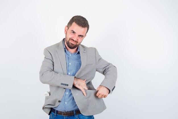 Jeune homme en chemise, jeans, veste de costume, ouverture de la poche de la veste avec les mains et à la joyeuse, vue de face.