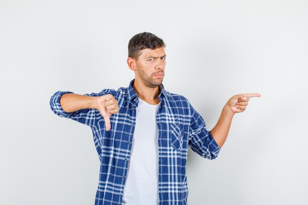 Jeune homme en chemise pointant vers le bas avec le pouce vers le bas et regardant mécontent, vue de face.