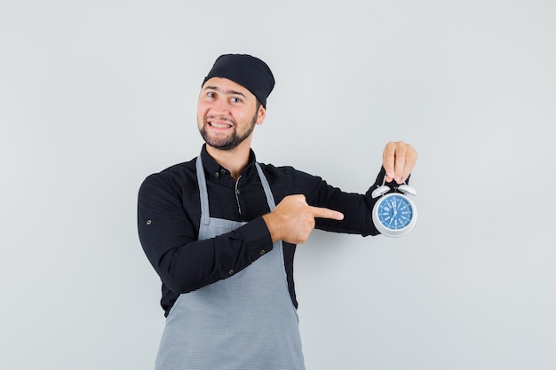 Jeune homme en chemise, tablier pointant sur le réveil et à la joyeuse vue de face.