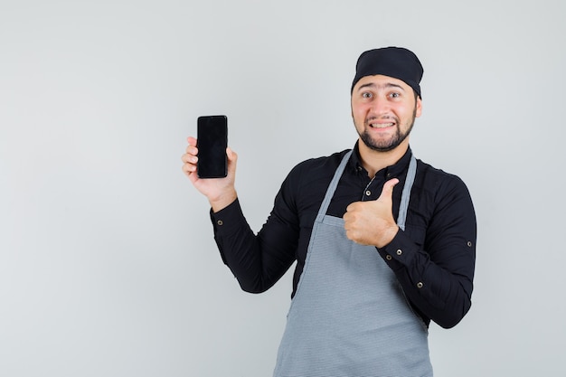 Jeune homme en chemise, tablier tenant le téléphone mobile avec le pouce vers le haut et à la vue de face, heureux.