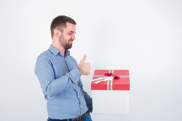 Jeune homme en chemise tenant une boîte-cadeau tout en montrant le pouce vers le haut et à la vue de face, heureux.