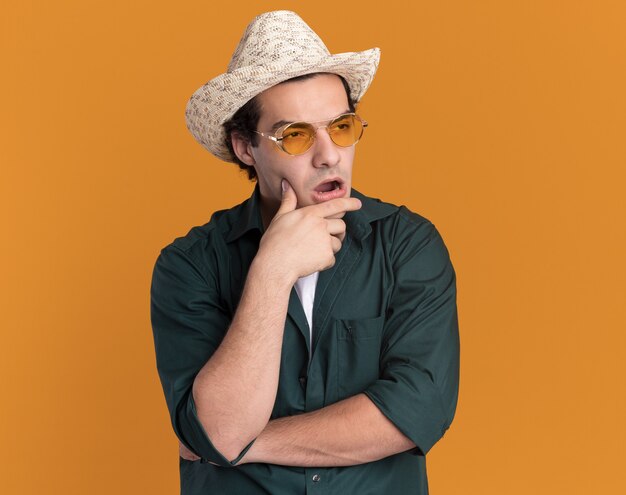 Jeune homme en chemise verte et chapeau d'été portant des lunettes à côté perplexe debout sur le mur orange