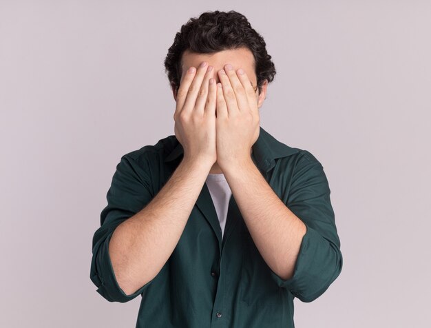 Jeune homme en chemise verte fermant les yeux avec la main debout sur un mur blanc