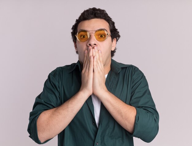 Jeune homme en chemise verte portant des lunettes à l'avant d'être choqué couvrant la bouche avec les mains debout sur un mur blanc
