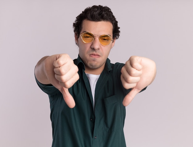 Jeune homme en chemise verte portant des lunettes à l'avant d'être mécontent montrant les pouces vers le bas debout sur un mur blanc