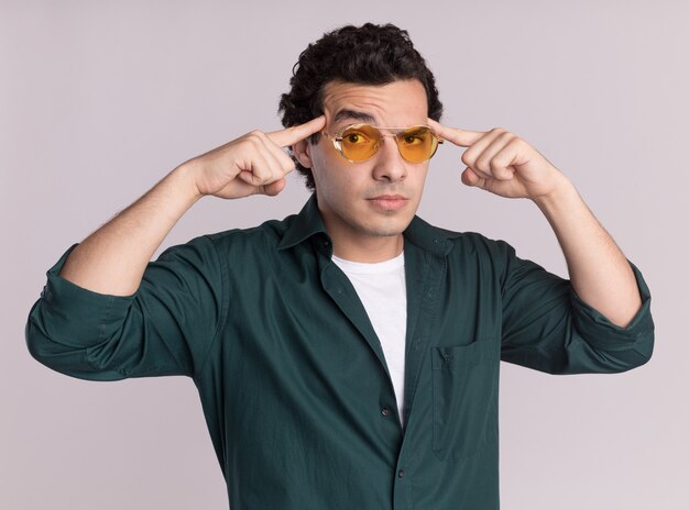 Jeune homme en chemise verte portant des lunettes à l'avant avec une expression confiante sur le visage intelligent pointant avec l'index sur ses tempes debout sur un mur blanc