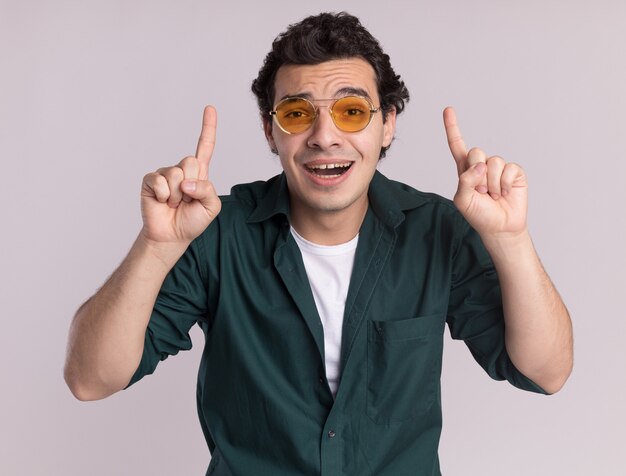 Jeune homme en chemise verte portant des lunettes à l'avant heureux et surpris montrant l'index debout sur un mur blanc