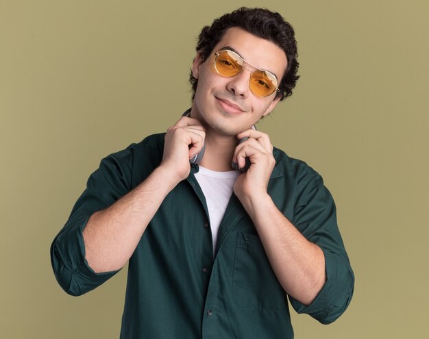 Jeune homme en chemise verte portant des lunettes à l'avant souriant confiant debout sur mur vert