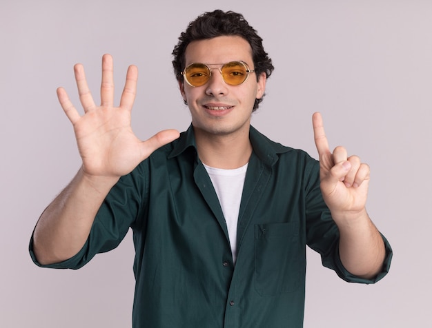 Photo gratuite jeune homme en chemise verte portant des lunettes à l'avant souriant confiant montrant numéro six debout sur un mur blanc