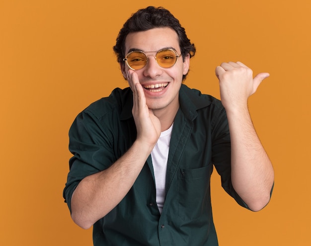 Jeune homme en chemise verte portant des lunettes à l'avant souriant joyeusement avec la main près de la bouche pointant avec le pouce sur le côté debout sur le mur orange