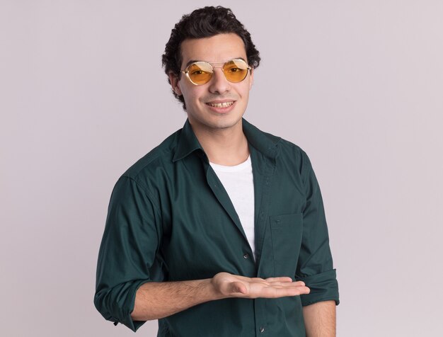 Jeune homme en chemise verte portant des lunettes à l'avant avec le sourire sur le visage présentant quelque chose avec le bras de la main debout sur un mur blanc