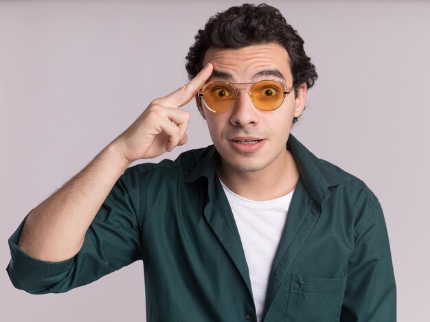 Jeune homme en chemise verte portant des lunettes à l'avant surpris en pointant avec l'index sur son temple ayant une nouvelle idée debout sur un mur blanc