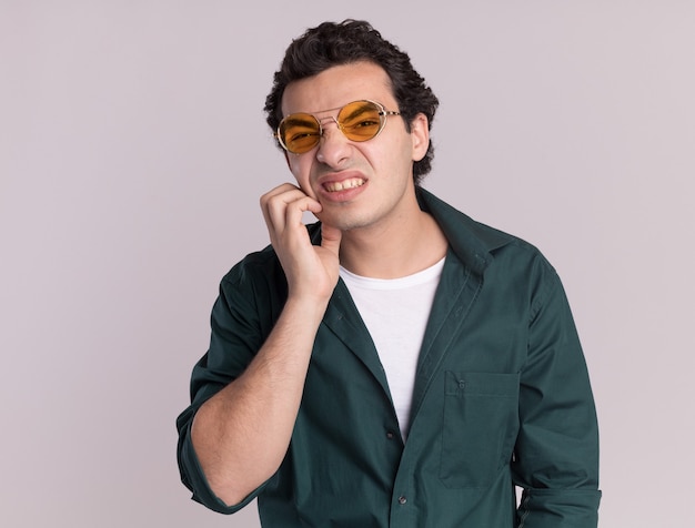 Jeune homme en chemise verte portant des lunettes à la confusion de se gratter le visage debout sur un mur blanc