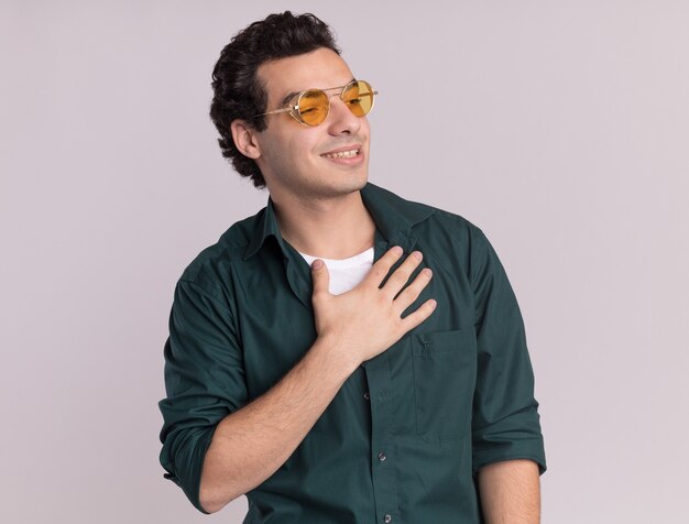 Jeune homme en chemise verte portant des lunettes à côté tenant la main sur sa poitrine souriant sentiment reconnaissant debout sur un mur blanc