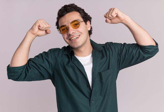 Jeune homme en chemise verte portant des lunettes heureux et excité en levant les poings célébrant la victoire debout sur un mur blanc