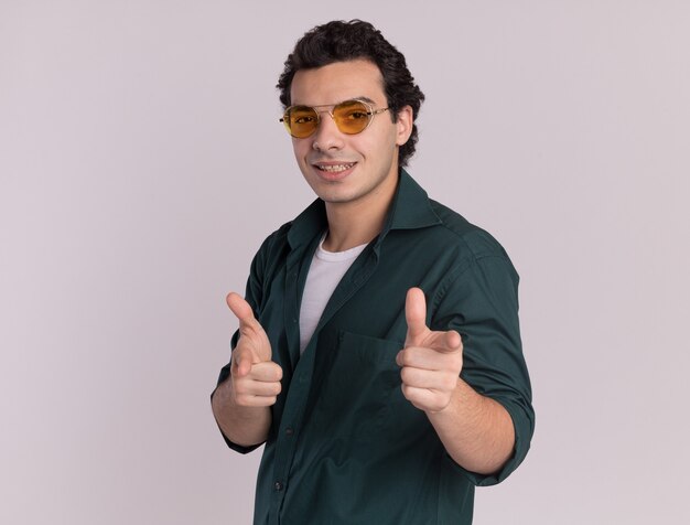 Jeune homme en chemise verte portant des lunettes pointant avec l'index à l'avant debout heureux et positif sur mur blanc