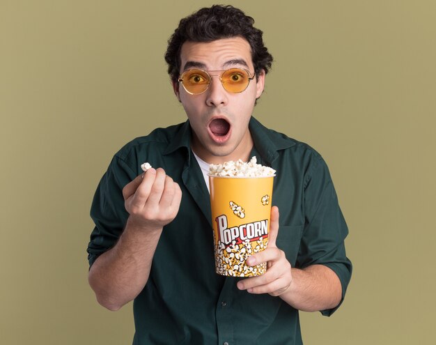 Jeune homme en chemise verte portant des lunettes tenant un seau avec du pop-corn à l'avant étonné et surpris debout sur le mur vert