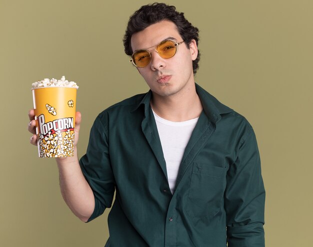 Jeune homme en chemise verte portant des lunettes tenant un seau avec du pop-corn à l'avant avec un visage sérieux debout sur un mur vert