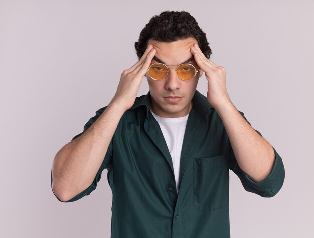 Jeune homme en chemise verte portant des lunettes touchant la tête fatiguée et surmenée ayant des maux de tête debout sur un mur blanc