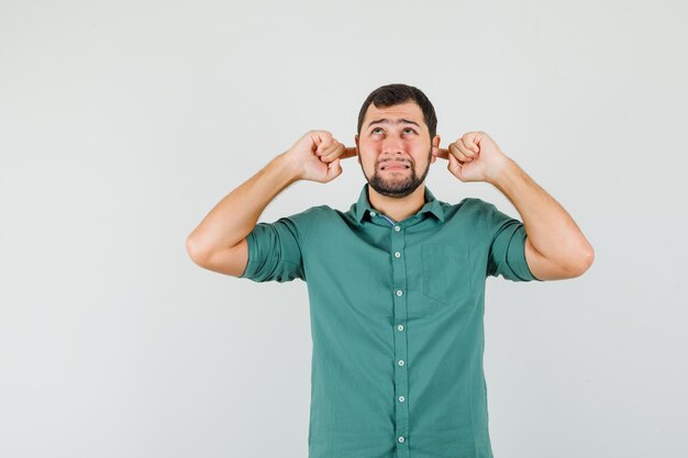 Jeune homme en chemise verte se bouchant les oreilles avec les doigts et ayant l'air effrayé, vue de face.