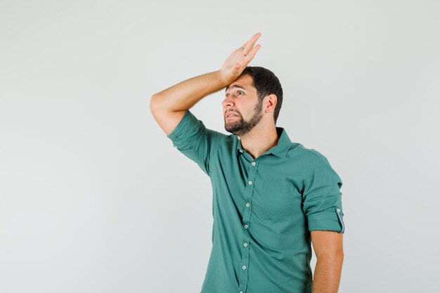 Jeune homme en chemise verte tenant la main sur la tête et l'air ennuyé, vue de face.