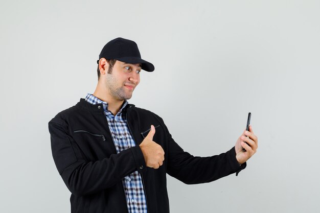 Jeune homme en chemise, veste, casquette montrant le pouce vers le haut tout en prenant selfie et à la joie