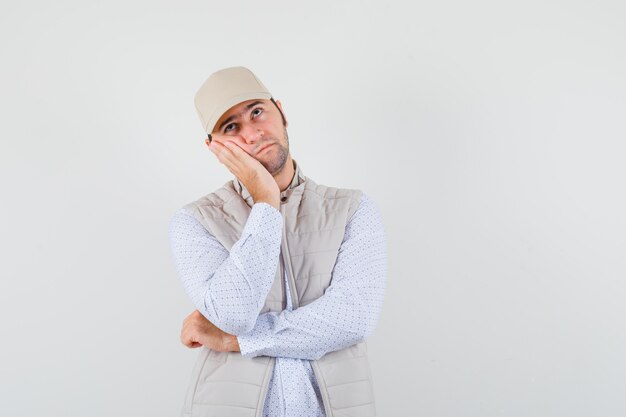 Jeune homme en chemise, veste sans manches, casquette appuyée sur sa paume tout en pensant et regardant pensif, vue de face.