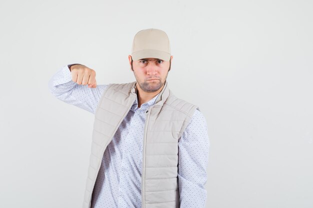 Jeune homme en chemise, veste sans manches, casquette levant son poing et regardant agressif, vue de face.