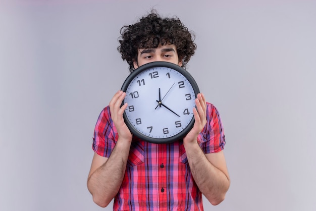 Jeune homme cheveux bouclés chemise colorée isolée se cachant derrière l'horloge