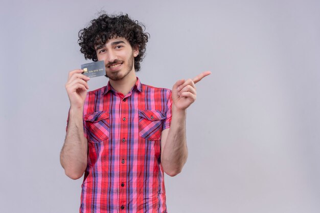 Jeune homme cheveux bouclés isolé carte bancaire chemise colorée souriant et pointant