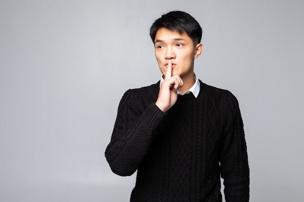 Jeune homme chinois asiatique demandant de se taire avec le doigt sur les lèvres, debout sur un mur blanc isolé. Silence et concept secret.