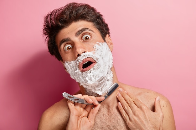 Un jeune homme choqué applique de la mousse, se prépare à tailler la barbe, tient la lame de rasoir, se sent épais et fatigué du rasage quotidien