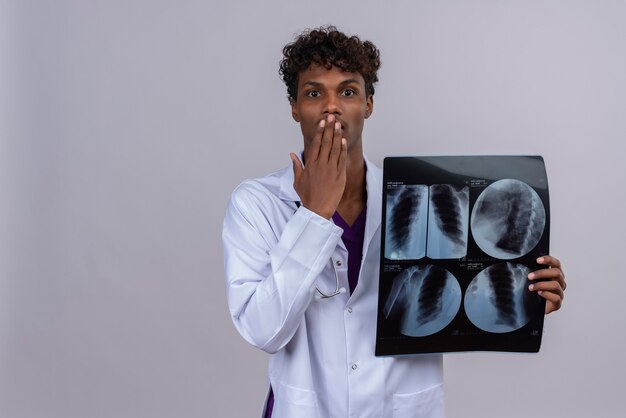 Un jeune homme choqué beau médecin à la peau sombre avec des cheveux bouclés portant un blouse blanche avec un stéthoscope montrant un rapport de rayons x