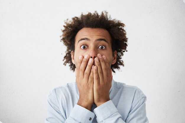 Jeune homme avec une coiffure afro couvrant la bouche et regardant la caméra avec une expression effrayée sur son visage