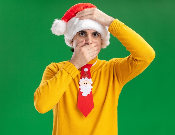 Photo gratuit jeune homme en col roulé jaune et bonnet de noel avec cravate drôle avec la main sur sa tête couvrant la bouche avec la main debout sur le mur vert