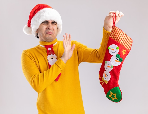 Jeune homme à col roulé jaune et bonnet de noel avec cravate drôle tenant bas de Noël en le regardant avec une expression dégoûtée debout sur fond blanc